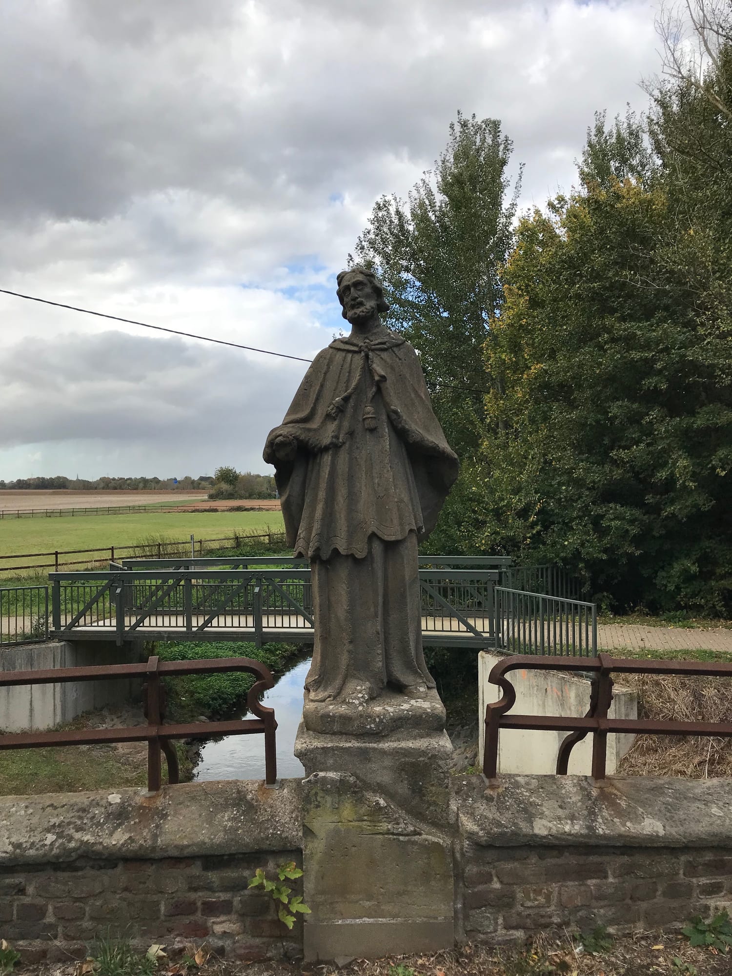 Stone statue of a robed figure set against the rural landscape of Gymnich with open fields in the background
